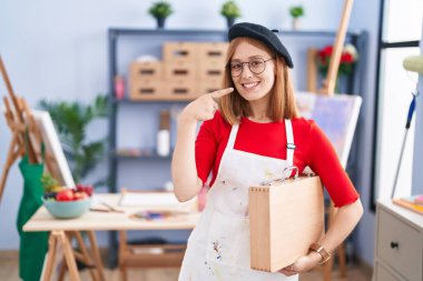 Sanat stüdyosundaki genç kızıl saçlı kadın elinde sanat çantası, gülümsüyor ve parmaklarıyla ve ağzıyla işaret ediyor. diş sağlığı kavramı. 