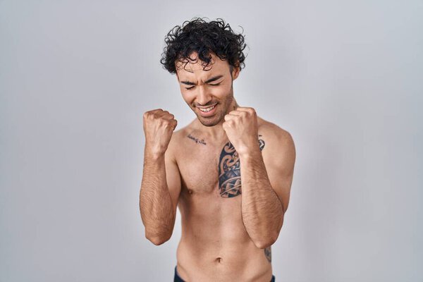 Латиноамериканец стоя без рубашки очень счастлив и взволнован делая жест победителя с поднятыми руками, улыбаясь и крича об успехе. празднование концепции. 