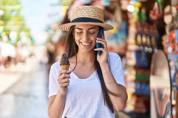 Young hispanic woman tourist talking on the smartphone eating ice cream at street market