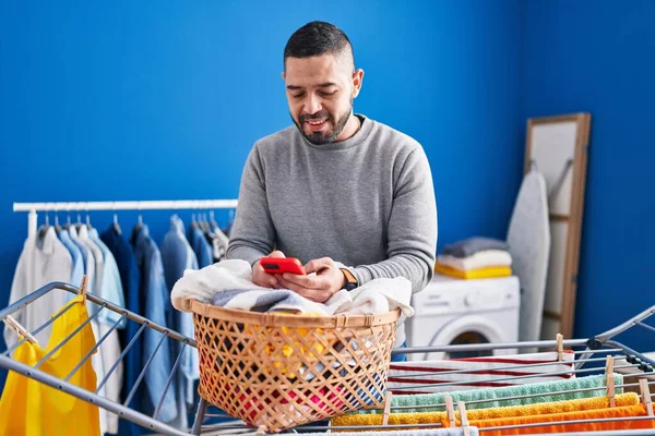 年轻的拉丁男人在洗衣房用智能手机把衣服挂在晾衣绳上 — 图库照片