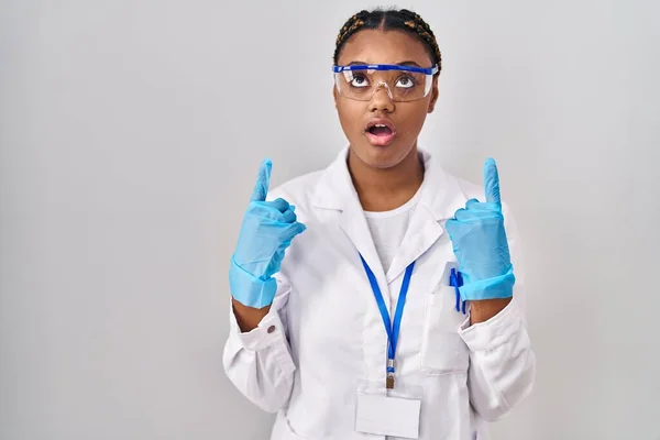 身穿科学家长袍 头戴辫子的非洲裔美国妇女惊奇而惊奇地抬起头 用手指和胳膊指指着 — 图库照片