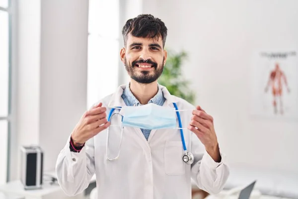 Νεαρός Ισπανός Γιατρός Γενειάδα Που Κρατάει Μάσκα Ασφαλείας Χαμογελώντας Ένα — Φωτογραφία Αρχείου