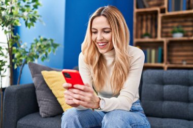 Evdeki kanepede oturan akıllı telefon kullanan genç sarışın kadın.