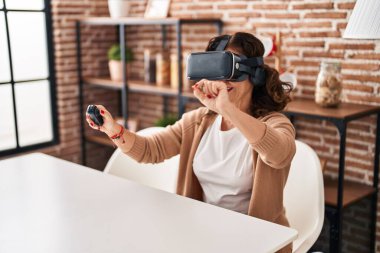 Middle age hispanic woman wearing virtual reality glasses at home