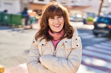 Orta yaşlı kadın gülümsüyor. Kollarını kavuşturup caddede duruyor.