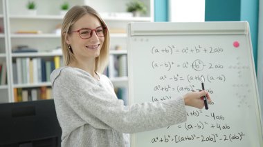 Young blonde woman teaching maths on magnetic board at library university