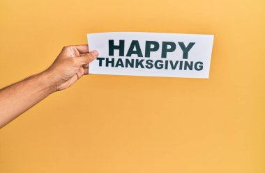 Hand of caucasian man holding paper with happy thanksgiving message over isolated yellow background