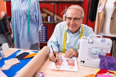 Orta yaşlı, gri saçlı, sırıtan, kendine güvenen, atölyede kıyafet tasarımı çizen bir adam.