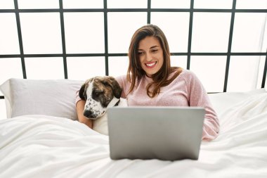 Yatak odasında dizüstü bilgisayar kullanan, köpeğini kucaklayan genç bir kadın.