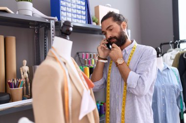 Young hispanic man tailor talking on smartphone standing by manikin at atelier