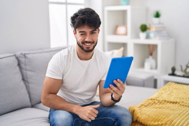 Sakallı İspanyol bir adam kanepeye oturmuş kameraya bakıyor. Seksi bir ifadeyle, neşeli ve mutlu bir yüzle.. 