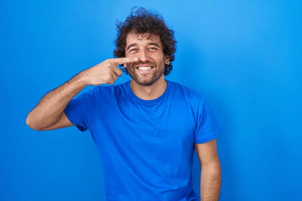ヒスパニック系の若い男の青の背景に顔と鼻に手の指を指して立って 陽気な笑顔 美の概念 — ストック写真