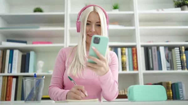 Junge Blonde Studentin Schreibt Mit Smartphone Und Kopfhörer Auf Notizbuch — Stockfoto