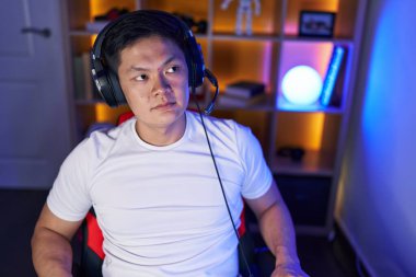Young chinese man streamer sitting on table with serious expression at gaming room