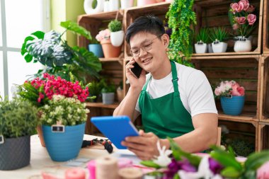 Young chinese man florist talking on smartphone using touchpad at flower shop