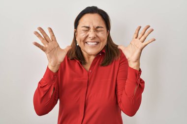 Beyaz arka planda dikilen Latin kökenli olgun bir kadın başarının çılgınlığını kollarını kaldırarak ve gözlerini kapayarak kutluyor. Heyecanlı çığlık atıyor. Kazanan konsept 