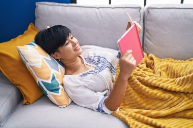 Orta yaşlı Çinli kadın evdeki kanepede uzanmış kitap okuyor.