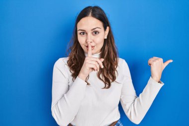 Young hispanic woman standing over blue background asking to be quiet with finger on lips pointing with hand to the side. silence and secret concept. 