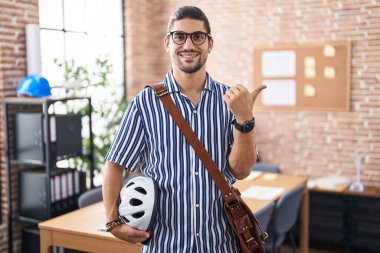 Ofiste çalışan uzun saçlı İspanyol bir adam bisiklet kaskı tutuyor. Arka tarafı işaret ediyor. El ve baş parmakları havada, kendinden emin gülümsüyor. 