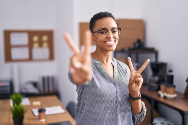 Ofiste çalışan, gülümseyen gözlüklü, parmaklarını zafer işareti yaparken gösteren Afrikalı Amerikalı bir kadın. İki numara.. 