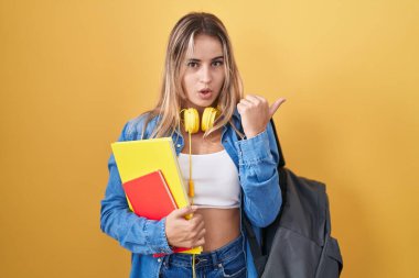 Sırt çantası takan genç sarışın bir kadın ve elinde kitaplar var. Şaşırmış bir şekilde el ile işaret ediyor, ağzı açık ve şaşırmış bir şekilde ifade ediyor.. 