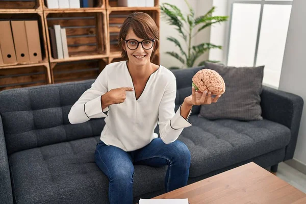 Brunette woman working at therapy office holding brain pointing finger to one self smiling happy and proud