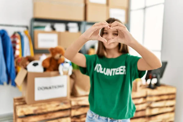 Adorable girl wearing volunteer uniform doing heart symbol with hands at charity center