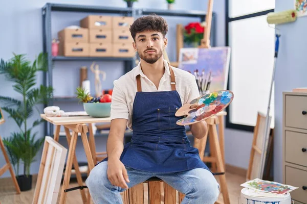 Sakallı Arap bir adam sanat stüdyosunda oturuyor paleti tutuyor. Duruşunu düşünüyor ve kendine güveni tam görünüyor. 