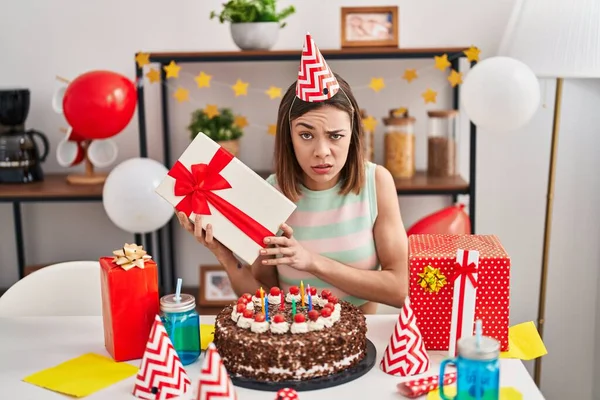 Hispanic woman celebrating birthday with cake holding gift skeptic and nervous, frowning upset because of problem. negative person.