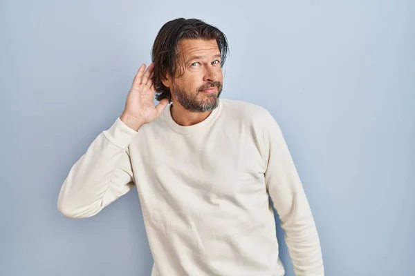 噂やゴシップに耳を傾ける耳の上に手で笑みを浮かべて青い背景にカジュアルなセーターを着てハンサムな中年の男性 聴覚障害の概念 — ストック写真