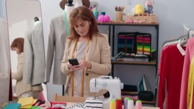 Orta yaşlı kadın terzi elbise fabrikasında akıllı telefon kullanarak gülümsüyor.
