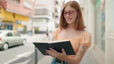 Young redhead woman wearing glasses reading book at street