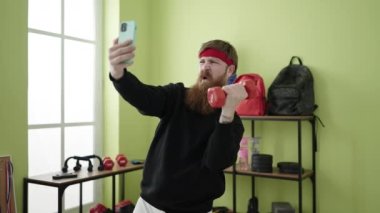 Genç kızıl saçlı adam spor merkezinde dambıl eğitimi alıyor ve akıllı telefonlardan selfie çekiyor.