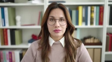 Young beautiful hispanic woman student sitting on table with i dont know expression at library university