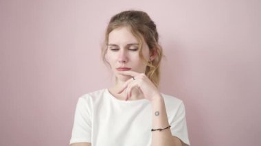 İzole edilmiş pembe arka planda şüpheli bir ifadeyle duran genç sarışın kadın.