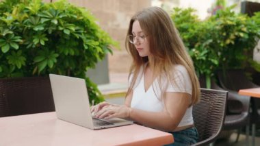 Genç sarışın kadın kahve terasındaki masada dizüstü bilgisayar kullanıyor.