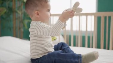 Sevimli çocuk yatakta oturmuş, ciddi bir ifadeyle yatak odasında oyuncaklarla oynuyor.