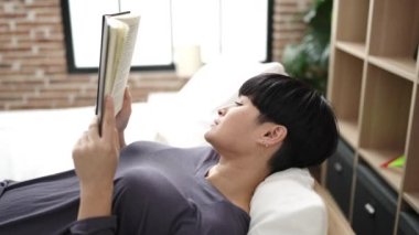 Young chinese woman reading book lying on bed at bedroom