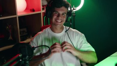 Young hispanic man streamer smiling confident doing heart symbol with hands at gaming room