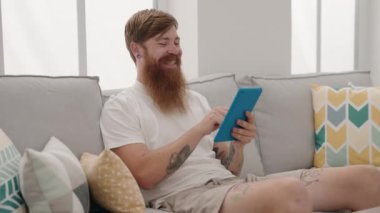 Young redhead man using touchpad sitting on sofa at home