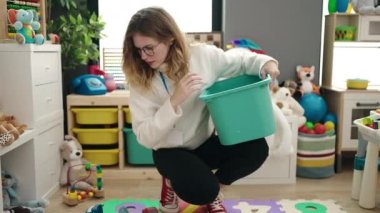 Young blonde woman preschool teacher picking up toys at kindergarten