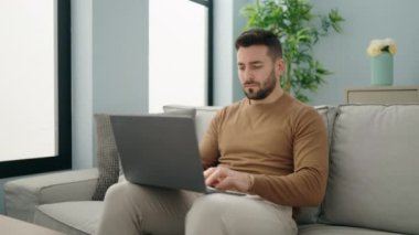 Young hispanic man using laptop sitting on sofa at home