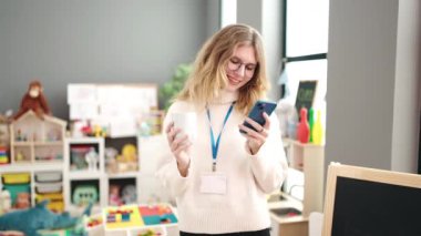 Young blonde woman preschool teacher using smartphone drinking coffee at kindergarten