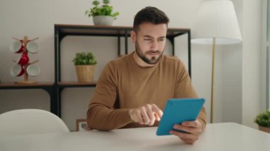 Young hispanic man using touchpad sitting on table at home