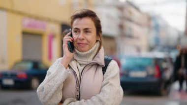 Orta yaşlı bir kadın gülümsüyor. Sokakta akıllı telefondan konuşuyor.