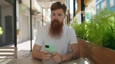 Young redhead man using smartphone with winner expression at coffee shop terrace