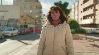 Orta yaşlı kızıl saçlı kadın gülümsüyor. Kollarını kavuşturup caddede duruyor.