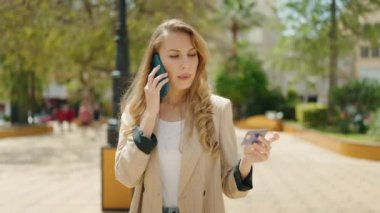 Genç sarışın kadın akıllı telefondan konuşuyor ve parkta kredi kartı kullanıyor.