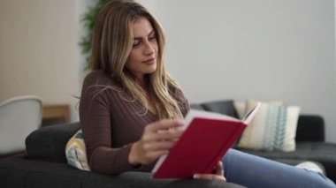 Young blonde woman reading book sitting on sofa at home