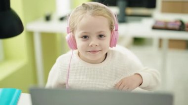Laptop ve kulaklık kullanan sevimli sarışın kız sınıfta baş parmağıyla hareket yapıyor.